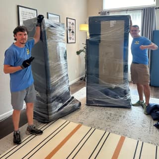 A couple of movers in blue shirts with KZ2 Moving Company logos carefully wrapping large pieces of furniture in protective padding inside a brightly lit living room, ensuring safe transport as part of a professional moving service.