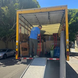 A mover in a blue shirt stands at the back of a yellow moving truck, organizing boxes and furniture for a residential move, ensuring efficient loading.