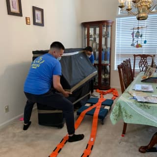 Two strong movers in a dining room use moving straps to secure a piano, ensuring safe transport of the heavy item during a residential move.