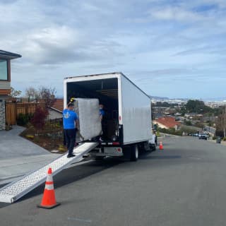 Two professional movers load a large wrapped with layers of shrink wrap mattress onto a moving truck parked on a sloped residential street, showcasing their skill in handling bulky items during a move.