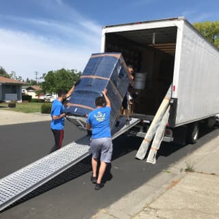 Three movers guide a large wrapped item up a ramp into a moving truck, demonstrating teamwork and safety in their moving process.