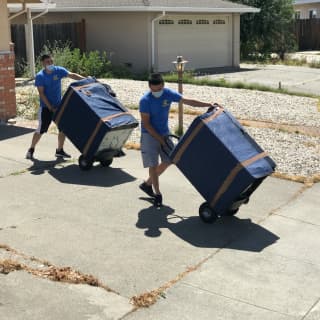 Two movers use dollies to transport wrapped washer and drier across a driveway, exemplifying efficiency in residential moving services.