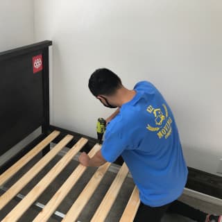 A moving expert uses a power drill to disassemble a bed frame in a bedroom, preparing furniture for safe transport during a residential move.