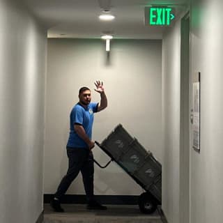 A busy mover waves while pushing a stack of heavy storage bins on a dolly through a narrow hallway, emphasizing friendly service and efficient moving.