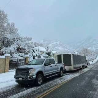 A truck with a long moving trailer drives through a snowy mountain pass, braving challenging weather conditions to deliver professional moving services, even in winter.