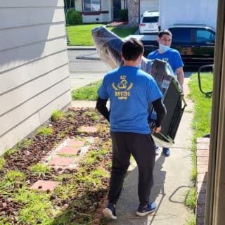 Two movers in face masks carry a large, padded item up a front yard path, showcasing their strength and careful handling during a residential move.